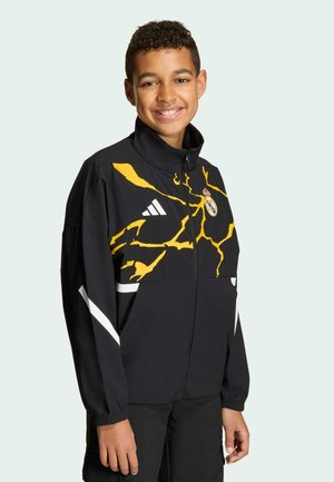 Czarna kurtka z żółtym wzorem błyskawicy, białymi paskami, zamek błyskawiczny i wysoki kołnierz. Na piersi znajduje się logo Realu Madryt.