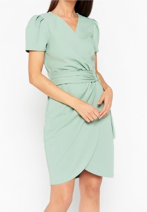 Kobieta w jasnozielonej sukience z falbanami i krótkimi puffowanymi rękawami, z szerokim paskiem z złotą klamrą na białym tle.