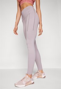 Leggings violet clair avec taille haute, texture lisse et poches latérales. Portés avec des chaussures de sport rose clair avec des détails en mesh.
