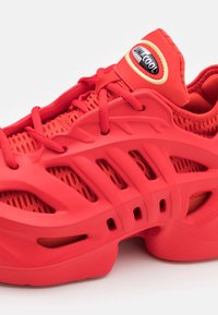 Červené sportovní boty mají síťovaný svršek, tlustou gumovou podrážku a výrazné logo značky na polstrovaném jazyku, což představuje moderní design.