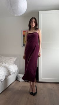 Jonge vrouw staand binnen, gekleed in een mouwloze pruimkleurige midi-jurk en zwarte hoge hakken in een minimalistische slaapkamer.