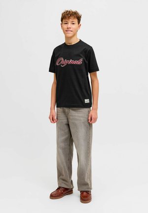 Tinejdžer s kovrčavom kosom koji nosi crnu majicu s natpisom "Originals", sive široke traperice i smeđe cipele, stojeći nasuprot jednostavne bijele pozadine.