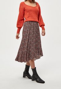 Roter Strickpullover mit Ballonärmeln, kombiniert mit einem gemusterten, ausgestellten knöchellangen Rock mit Blumenmotiven und schwarzen Knöchelstiefeln.