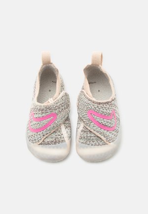 Para dziecięcych dzianinowych trampek w odcieniach beżu i brązu, z różowymi logo Nike w kształcie łyżwy oraz pętelkami ułatwiającymi zakładanie, widziane z góry na białym tle.