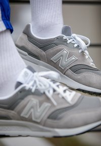 Paire de baskets grises New Balance avec des lacets blancs portées avec des chaussettes blanches, affichées en gros plan sur un fond neutre.
