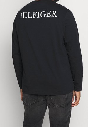 Personne portant un t-shirt noir à manches longues avec l'inscription "HILFIGER" en lettres blanches sur le haut du dos et un jean gris foncé, debout, de dos.