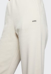 Helle beige Jogginghose aus weichem Baumwollmix, mit einer Seitentasche und einem kleinen schwarzen 'JJXX'-Logo auf dem Oberschenkel. Glatte Textur.