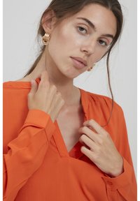 Orange Bluse mit langen Ärmeln und V-Ausschnitt, mit glatter Textur. Goldene Creolen sind zu sehen. Das Model berührt mit einer Hand den Kragen.