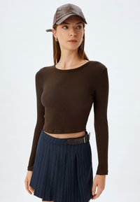 Brązowy, prążkowany, długi rękaw crop top w połączeniu z granatową, prążkowaną, plisowaną spódnicą z paskiem, noszoną z brązową skórzaną czapką.
