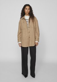 Braune Wollmischung, oversized Jacke mit Knopfverschluss, zwei Brusttaschen und geradem Schnitt, kombiniert mit schwarz-weit geschnittenen Hosen.