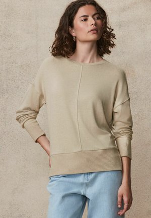 Pull en tricot beige clair avec un col rond, des poignets et un ourlet côtelés. Présente un design fendu à l'avant et une texture douce.