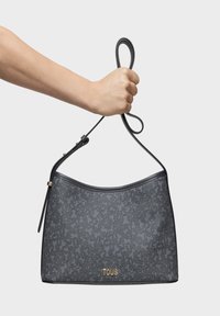 Bolso de hombro texturizado en gris con un diseño curvo en la parte superior, acentos de cuero y un logo dorado de "TOUS" en la parte inferior. Mano sosteniendo el bolso.