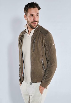 Brown suede bomber jacket with ribbed cuffs, zippered pockets, and a textured design. Worn over a light-colored polo shirt, model stands against a gray background.