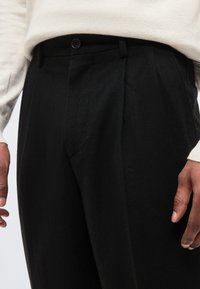 Μαύρο παντελόνι με ψηλή μέση, θηλιές για ζώνη, εμπρόσθιο κουμπί και φερμουάρ, συνδυασμένο με κρεμ μακρυμάνικη μπλούζα, σε κοντινή εικόνα που δείχνει γοφούς και χέρια.