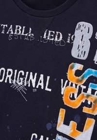 T-shirt en coton bleu marine arborant des textes blancs, orange et bleu avec diverses polices et des détails de couture, y compris l'année "1988".