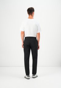Black trousers with an elastic waistband, paired with a white short-sleeve shirt. The model wears gray slip-on shoes, viewed from the back.