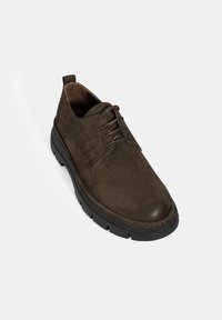Chaussure en daim marron foncé avec un bout rond, un laçage et une épaisse semelle en caoutchouc noir dotée de motifs de traction pour une meilleure adhérence.