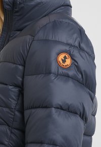 Doudoune bleu marine avec un design matelassé, une texture lisse, et un patch circulaire orange avec un logo de canard sur l'épaule.