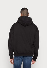 Felpa nera con cappuccio a vestibilità ampia, caratterizzata da un grande cappuccio, polsini e orlo a coste, realizzata in un tessuto morbido, indossata con jeans blu.