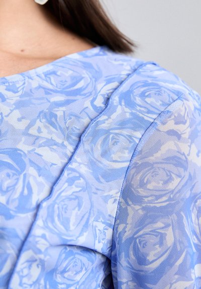 Ανοιχτό μπλε μπλούζα με λουλουδάτο μοτίβο τριαντάφυλλων. Το υλικό είναι ημι-διάφανο με υφή, ολοκληρωμένο με στρογγυλή λαιμόκοψη.