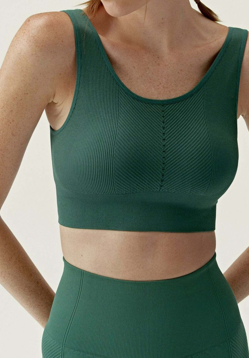 Sportovní podprsenka v lesní zelené barvě s ribkovým vzorem, s kulatým výstřihem, širokými ramínky a jemnými liniemi podél poprsí, které poskytují přiléhavou strukturu.