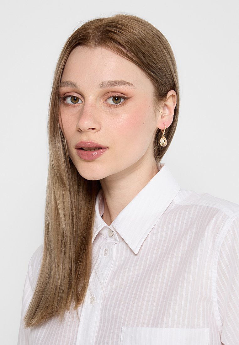Weißes Hemd mit vertikalen Streifen, mit Kragen und Tasche. Das Modell hat lange glatte Haare und trägt einen tropfenförmigen Ohrring.