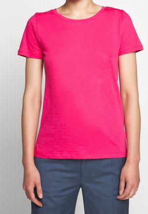 Persoon die een felroze korte mouwen T-shirt en donkerblauwe broek draagt, staand met ontspannen armen langs het lichaam.