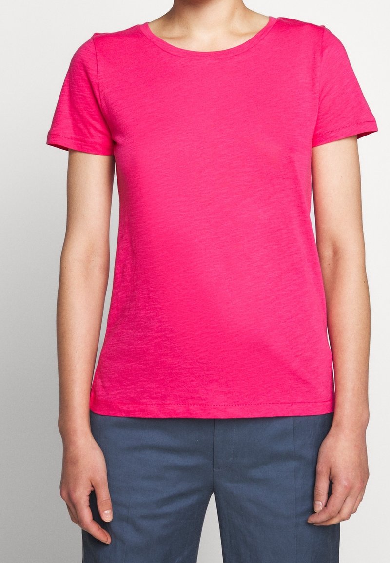 Personne portant un T-shirt court rose vif et un pantalon bleu foncé, debout avec les bras détendus le long du corps.