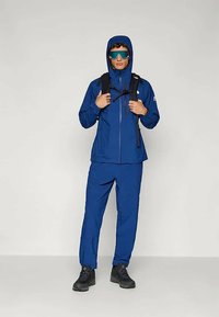 Μπλε αδιάβροχο μπουφάν με κουκούλα, συνδυασμένο με ταιριαστά παντελόνια. Διαθέτει φερμουάρ μπροστά, τσέπες και σακίδιο. Φοριέται με γυαλιά ηλίου και μπότες.