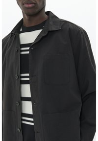 Black button-up jacket with a pointed collar and a chest pocket, worn over a black and white striped sweater. Smooth texture, casual design.