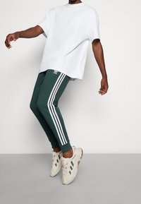 Osoba v tmavě zelených teplácích Adidas s bílými pruhy a bílými teniskami, pózující s nohama ohnutýma a pažemi mírně nataženými.