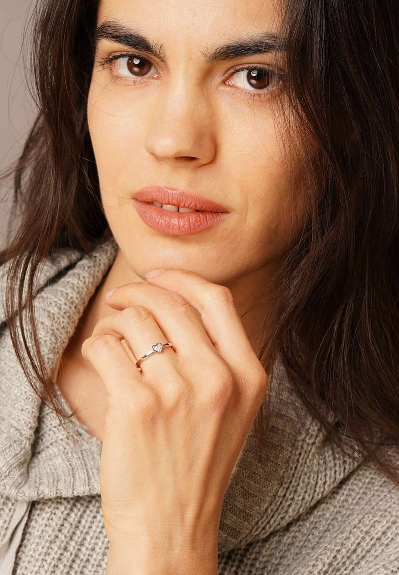 Goldring mit einem herzförmigen Stein, vor einem strukturierten grauen Pullover. Die Hand ist nachdenklich nahe dem Gesicht positioniert, was den Ring hervorhebt.