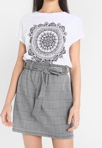 Camiseta blanca de algodón con estampado de mandala negra, combinada con una falda de talle alto a cuadros negros y blancos con detalle de cinturón anudado.