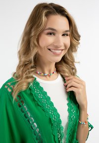 Zeleni kimono z čipkastim obrobom čez belo majico, dodatki vključujejo barvit obesek iz perlic in zapestnico z majhnim zvezdastim charmom.