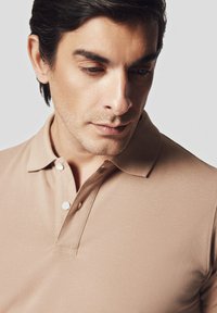 Beżowa koszulka polo z klasycznym kołnierzykiem, trzema guzikami, teksturowanym materiałem i dopasowanym krojem. Skupienie na okolicy szyi z zbliżeniem na szczegóły.