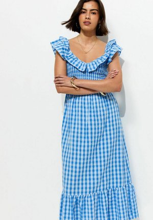 Mujer con cabello corto y oscuro con un vestido de cuadros azules y blancos con mangas con volantes, de pie con los brazos cruzados frente a un fondo blanco.