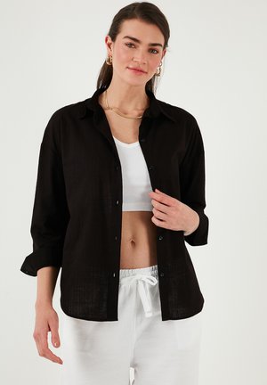 Black linen shirt with a relaxed fit, button-down front, and rolled-up sleeves, layered over a white ribbed top. White drawstring pants.
