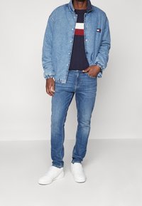 Denimová bunda s knoflíkovým zapínáním, světle modré barvy, elastické manžety; nošena přes námořnický svetr s bílými a  červenými pruhy, v kombinaci s modrými džíny a bílými teniskami.