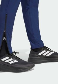 Czarne buty sportowe z białym wzorem trzech pasków; noszone z granatowymi spodniami z zamkami u dołu nogawek. Teksturowany materiał, lekka konstrukcja.