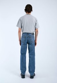 Camiseta de algodón gris y vaqueros de mezclilla azul. Los vaqueros tienen un corte recto, costuras visibles y detalles en los bolsillos traseros. Se llevan zapatos negros.