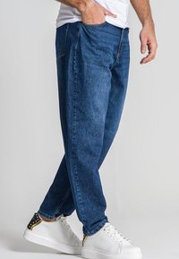 Vaqueros de mezclilla azul oscuro con un corte relajado, que cuentan con un diseño clásico de cinco bolsillos y sutiles detalles de costura. Llevados con zapatillas blancas.