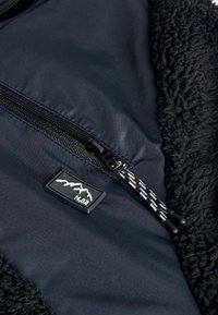 Czarna polarowa kurtka z detalami, posiadająca gładką podszewkę, kieszeń na zamek oraz naszywkę z logo w kształcie góry na teksturowanej powierzchni.