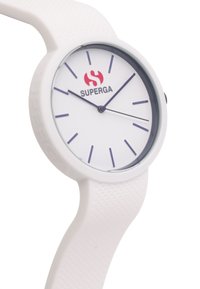 Reloj blanco con una cara redonda y marcadores de hora morados, que presenta una correa texturizada y flexible y un logo rojo en la esfera.
