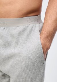 Γυμνό μέσο και χέρι ενός ατόμου που φοράει ανοιχτό γκρι παντελόνι με υφή και μπεζ ζώνη Calvin Klein, με το χέρι στην τσέπη.