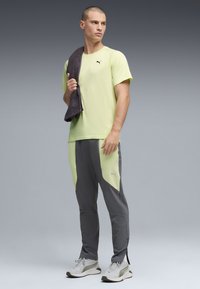 Άνδρας με κίτρινη μπλούζα Puma και γκρι-κίτρινο παντελόνι κρατώντας μαύρη πετσέτα στον ώμο του, φοράει λευκά αθλητικά Puma, στέκεται μπροστά σε γκρι φόντο.