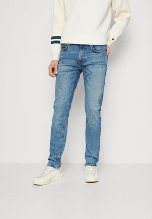 Mann trägt hellblaue Slim-Fit-Jeans, weiße Sneakers und einen weißen Pullover mit marineblauen Streifen an den Ärmelbündchen, steht vor weißem Hintergrund.