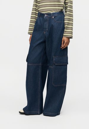 Személy laza sötétkék farmer cargo nadrágot visel nagy oldalzsebekkel és csíkos hosszú ujjú pólót, egy egyszerű háttér előtt áll.