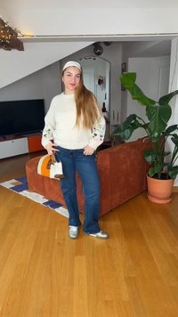 Jeune femme aux cheveux longs portant un pull blanc brodé, un jean bleu, des chaussures argentées et un bandeau, debout dans un salon.