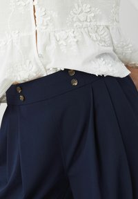 Blouse blanche brodée rentrée dans un pantalon marine taille haute avec des plis et quatre boutons visibles sur la ceinture.