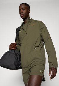Olivenfarget treningsjakke med glidelås og sidelomme, kombinert med matchende shorts. Svart treningsbag med glidelås, bæres i den ene hånden.